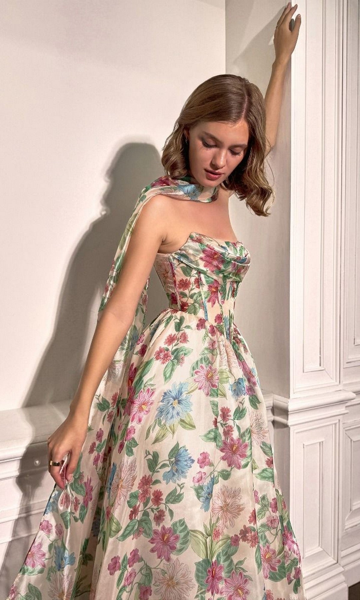 Wearing the Andrea and Leo A1425 long floral-print prom dress, a woman stands indoors by a white paneled wall, looking down with one arm raised. The floor-length gownโs colorful pattern brings elegance to her look.